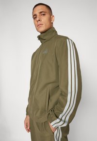Giacca leggera verde con colletto alto e design a tre strisce bianche sulle maniche. Presenta polsini elasticizzati e tasche laterali.