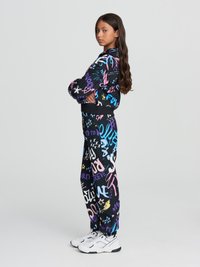 Tuta nera con motivi in stile graffiti multicolore, composta da una giacca con zip e pantaloni coordinati. Indossata con sneakers bianche da ginnastica.