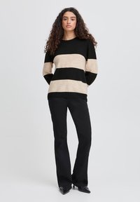 Suéter de rayas negras y beige con mangas largas, escote redondo, combinado con pantalones negros de campana y zapatos negros de punta, sobre un fondo neutro.