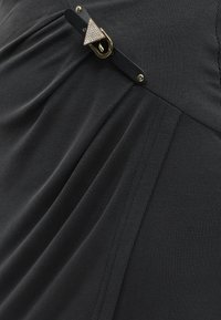 Tissu côtelé noir avec un détail froncé et une sangle noire avec une boucle triangulaire dorée ornée de petits strass.