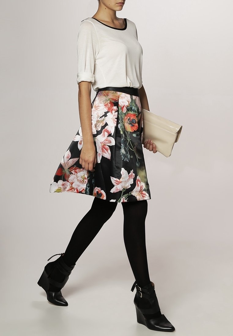 Femme marchant portant un chemisier blanc, une jupe fleurie mi-longue, des collants noirs, des bottines compensées noires et tenant une pochette beige.