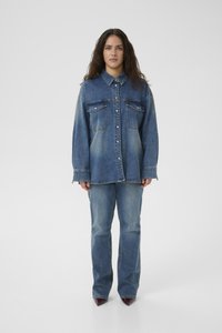 Camicia in denim oversize con chiusura a bottone, due tasche sul petto e maniche lunghe. Lavaggio blu chiaro a medio con aree leggermente sbiadite. Abbinata a jeans blu.