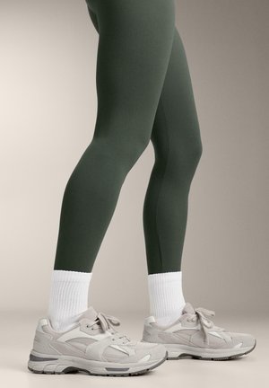 Picioare îmbrăcate în colanți verde închis, șosete albe până la gleznă și adidași activi gri deschis, stând pe un fundal neutru.