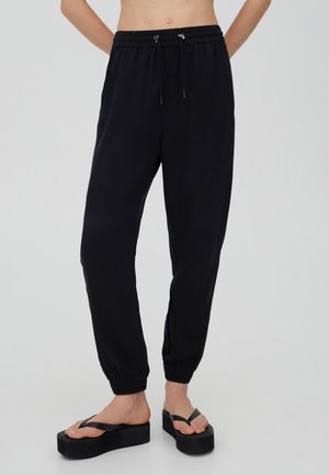 Pantalon de jogging noir à taille élastique avec cordon de serrage, chevilles resserrées, porté avec des sandales tongs plateformes noires sur une personne debout.