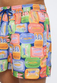 Κοντινό πλάνο πολύχρωμων μαγιό με σχέδιο από ετικέτες κονσερβοποιημένων ψαριών σε διάφορα χρώματα, δεμένα στη μέση με μπλε κορδόνι.