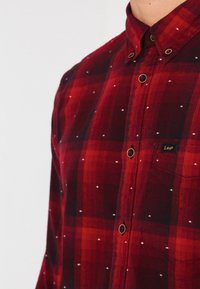 Camisa de cuadros rojos con botones, con cuadros contrastantes, cuello redondo y pequeños acentos de puntos blancos. Cuenta con un bolsillo en el pecho con marca y botones marrones.
