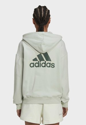 Personne aux cheveux tressés portant un sweat à capuche Adidas de couleur claire et un short assorti, vue de dos sur un fond uni.