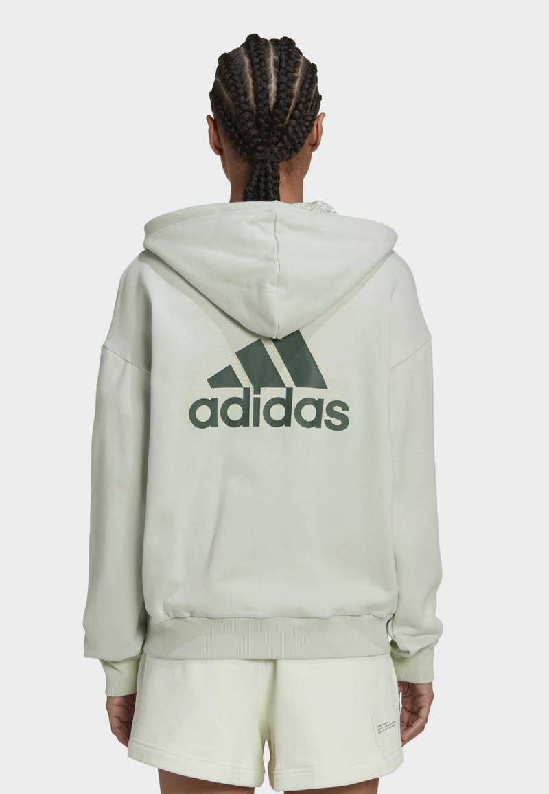 Personne aux cheveux tressés portant un sweat à capuche Adidas de couleur claire et un short assorti, vue de dos sur un fond uni.