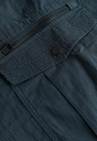 Dunkelblauer Stoff mit sichtbarem Reißverschluss und Knopfdetail, ausgestattet mit gestickten Mustern und glatter Textur, was auf ein strukturiertes Design hinweist.