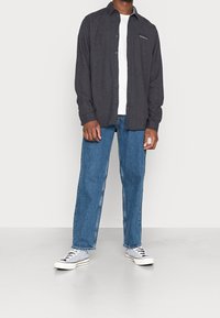 Ciemnoszara koszula zapinana na guziki na białym t-shircie, zestawiona z niebieskimi dżinsami i białymi trampkami w paski. Gładka tkanina i luźny krój.