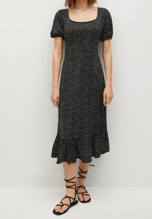 Femme portant une robe midi noire à pois blancs, manches bouffantes, encolure carrée et sandales plates noires à lacets.