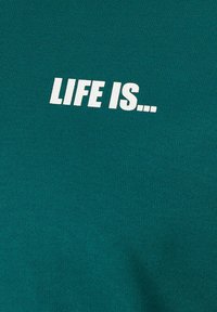 T-shirt in cotone teal con la scritta "LA VITA È..." in grassetto bianco. Il tessuto ha una texture liscia ed uniforme con scollatura a girocollo.