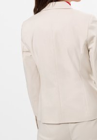 Jasnobeżowa, dopasowana marynarka o gładkiej fakturze, z usztywnionymi ramionami i pojedynczym rozcięciem z tyłu. W komplecie spodnie w tym samym kolorze.