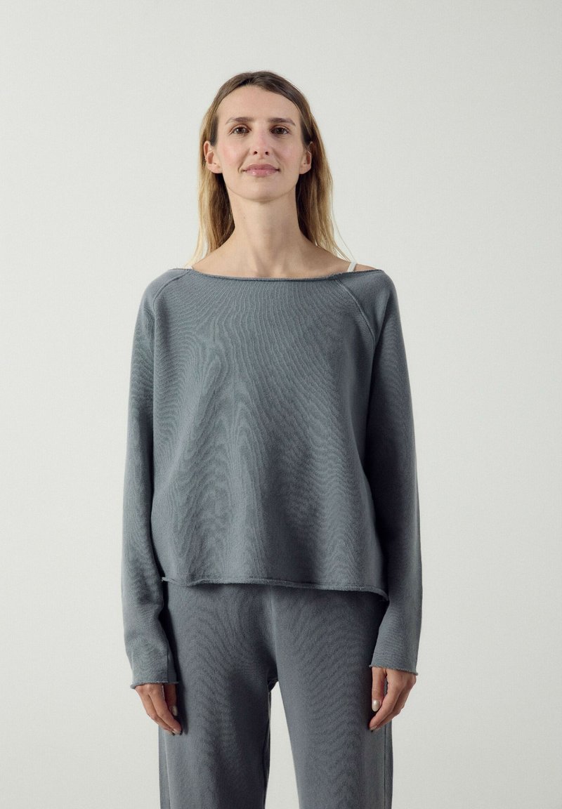Femme debout sur un fond clair, portant un haut gris ample à manches longues et un pantalon assorti avec une bretelle blanche visible sur son épaule gauche.
