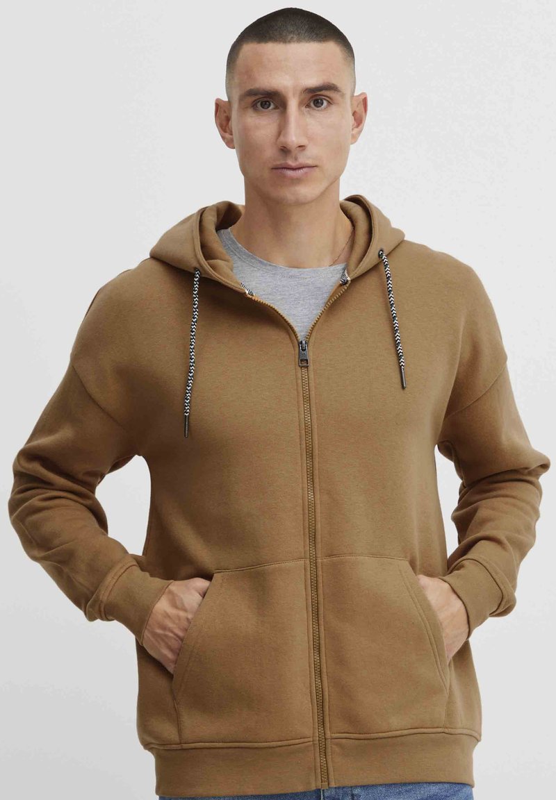 Hoodie zippé marron avec une capuche doublée, des cordons de serrage et des poches avant. Présente des poignets et un ourlet côtelés. Porté sur un t-shirt gris.