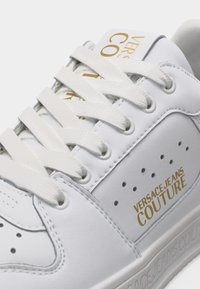 Λευκό πάνινο παπούτσι Versace Jeans Couture με λευκά κορδόνια, τρύπες αερισμού και χρυσό λογότυπο στο πλάι και στη γλώσσα.