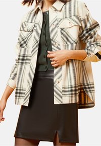 Kostkovaná bunda v krémové, zelené a černé barvě; má dvě náprsní kapsy, zapínání na knoflíky a dlouhé rukávy; spárovaná s černou sukní.