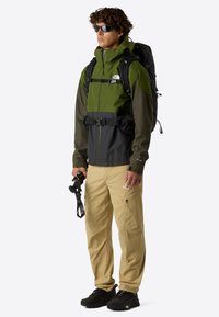 Giacca verde e grigia con zip frontale, cappuccio regolabile e cintura in vita. Pantaloni cargo beige con tasche con zip. Scarponi da trekking neri.