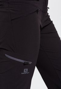 Svarta utomhusbyxor i lätt material. Har en dragkedjeficka på vänster lår och en logotyp på underbenet.