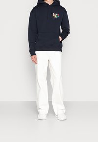 Sudadera marina con el texto "LOVE WINS" en letras coloridas, con un bolsillo frontal y cordones, combinada con pantalones anchos blancos y zapatillas blancas.