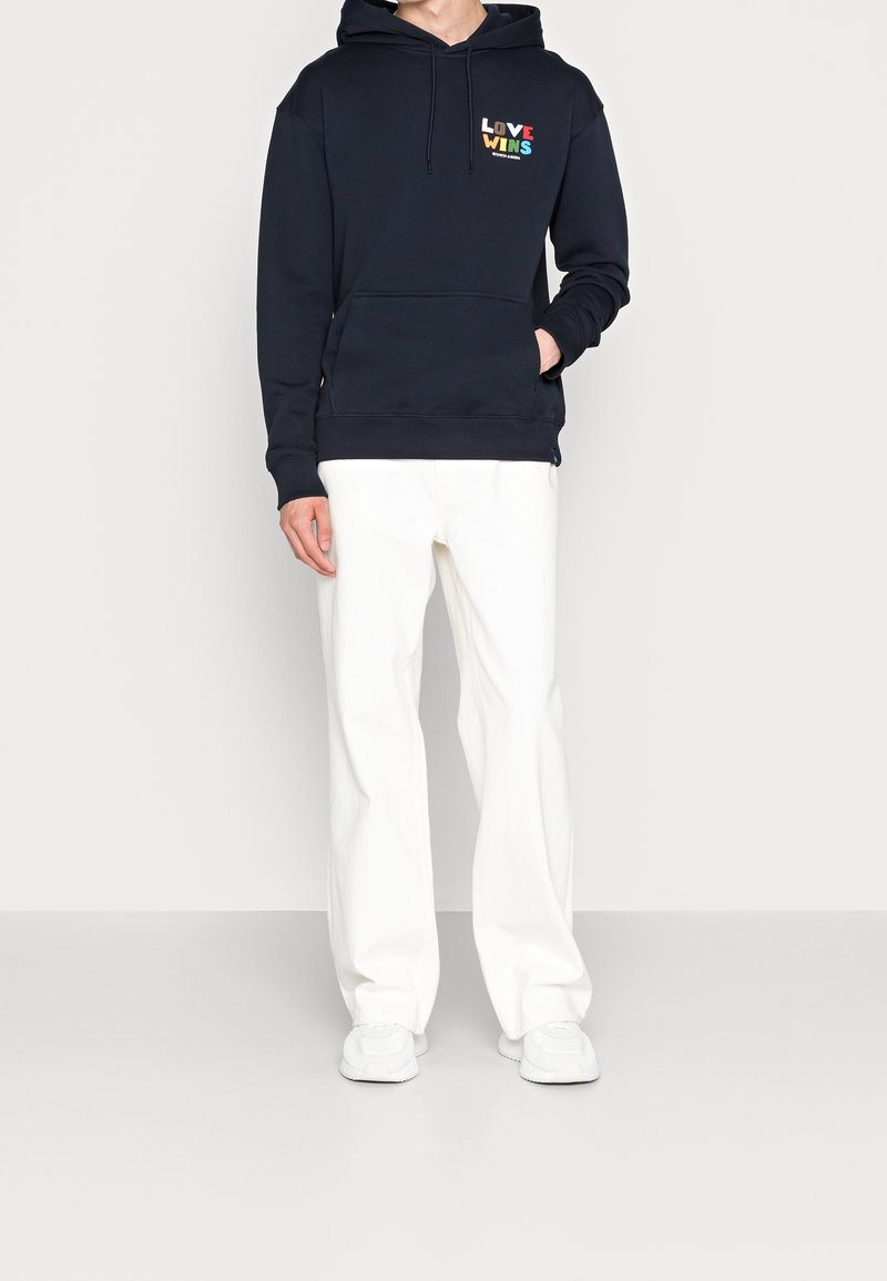 Marineblauwe hoodie met de tekst "LOVE WINS" in kleurrijke letters, voorzien van een voorzak en koordjes, gecombineerd met witte wijde broek en witte schoenen.