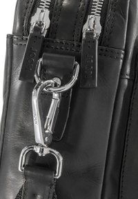 Dettaglio di una borsa di pelle nera con finiture in argento, dotata di doppie cerniere, di una tracolla in pelle con un gancio in cromo e di un marchio in rilievo.