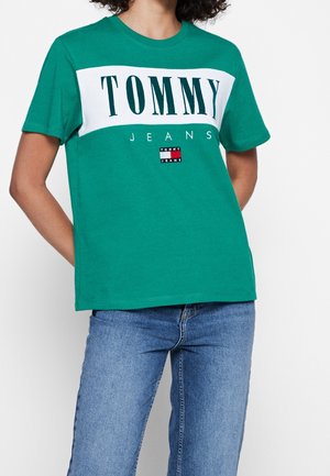 Person iført grøn Tommy Jeans T-shirt med hvidt logo og blå jeans, beskåret fra hals til midt på låret, står med hænderne bag ryggen.