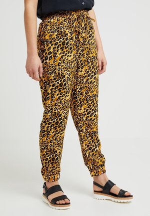 Frau trägt gelbe und schwarze Leopardenmusterhose mit elastischen Bündchen und schwarzen Plateausandalen vor schlichtem Hintergrund.