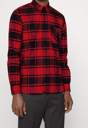 Hombre vistiendo una camisa de cuadros roja y negra con bolsillo en el pecho, combinada con pantalones gris oscuro, sobre un fondo liso.