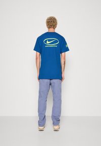 Modrá bavlněná trička s velkým bílým a neonovým logem na zadní straně a žlutým akcentem na rukávu, spárovaná s světle fialovými kalhotami.