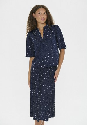 Marineblaues, kurzärmliges Kleid mit V-Ausschnitt und weißen Punkten. Glatter, leichter Stoff, der unter das Knie fließt und einen seitlichen Schlitz hat.