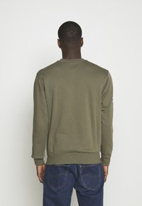 Hombre de espaldas, vistiendo un sudadera de manga larga color verde oliva y jeans azules, contra un fondo claro y liso.