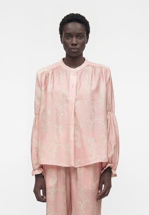 Femme aux cheveux courts et bouclés portant un chemisier rose ample et un pantalon assorti avec des motifs floraux clairs, debout sur fond blanc.