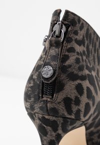 Zapato de tacón alto con estampado de leopardo negro y marrón, punta afilada, con cremallera lateral y detalle decorativo de botón metálico.