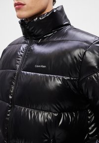 Μαύρο μπουφάν puffer με γυαλιστερή φινίρισμα, που διαθέτει οριζόντιες ραφές, ψηλό γιακά και κλείσιμο με φερμουάρ. Λογότυπο Calvin Klein στο στήθος.