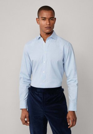 Camisa de botones azul claro con un patrón texturizado, mangas largas y un cuello clásico, combinada con pantalones oscuros de pana.