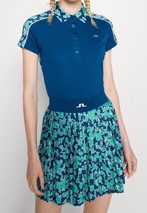 Frau trägt ein blaues Poloshirt mit floralem Kragen und Ärmeln, kombiniert mit einem plissierten Rock mit floralem Muster in Blau- und Grüntönen.