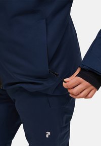 Person passt die Kordelzug auf einer marineblauen Jacke nahe der Hüfte an, trägt dazu passende Hosen mit einem Logo auf dem linken Oberschenkel und einem schwarzen Ärmelbündchen.