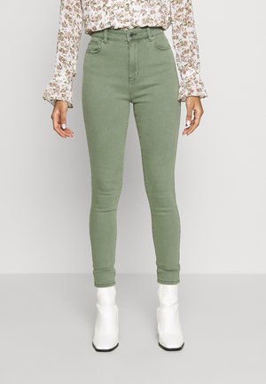 Kvinde iført højtaljede grønne skinny jeans, en langærmet blomstret bluse og hvide ankelstøvler, stående indendørs mod en neutral baggrund.