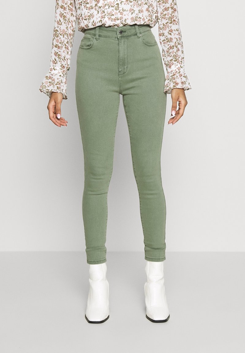 Femme portant un jean skinny vert taille haute, un chemisier fleuri à manches longues et des bottines blanches, debout à l'intérieur devant un fond uni.