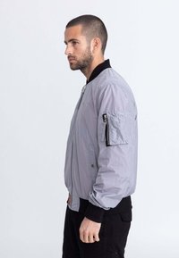 Bomber jacket grigio chiaro con colletto e polsini a coste neri, dotato di una tasca con zip sulla manica e una sottile texture. Chiusura frontale con zip.