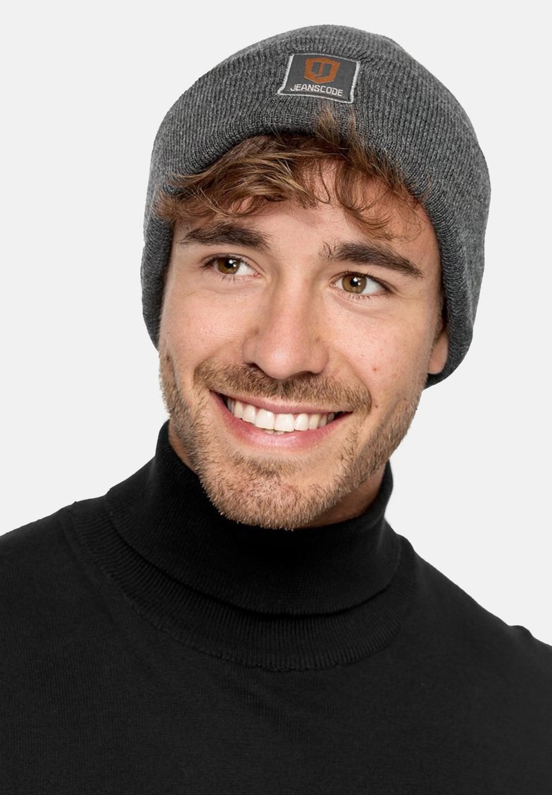 Gray knit beanie with a textured pattern and a branded patch. Worn with a black turtleneck. Simple and functional design.