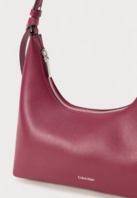 Burgundi színű bőr válltáska sima felülettel, ívelt formával, cipzáras záródással és ezüst színű hardverelemekkel, amelyek a Calvin Klein logóját viselik.