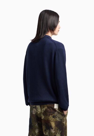 Maglione a coste blu navy con scollo rotondo e vestibilità ampia, abbinato a pantaloni multicolore a motivi testurizzati.