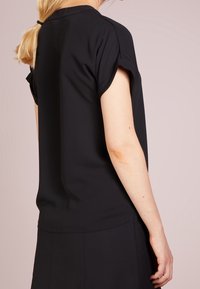 Kvinna med blont hästsvans som bär en svart kortärmad blus och svart kjol, sedd bakifrån mot en ljus beige bakgrund.