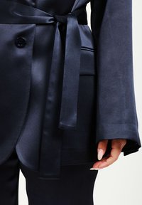 Blazer di raso blu navy con cintura, grande bottone frontale, maniche larghe e texture liscia, abbinato a pantaloni coordinati.