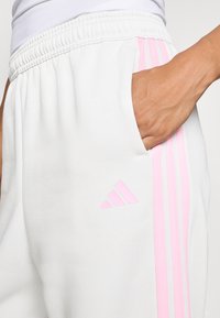Hånden hviler i lommen på hvide bukser med elastisk talje og rosa striber samt logo på siden.
