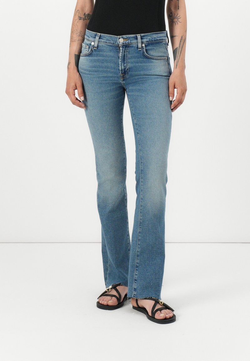 Jean flare bleu clair avec un délavage, cinq poches et une fermeture éclair. Accompagné de sandales plates noires avec un accent doré.