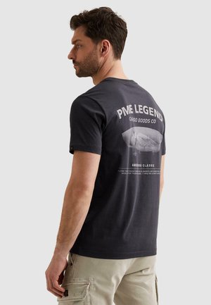 Mężczyzna z brodą, ubrany w czarną koszulkę PME Legend z nadrukiem oraz beżowe szorty cargo, patrzący w lewo na jednolitym tle.