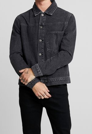 Person iført en mørkegrå denimjakke med knapper og brystlomme, sorte bukser og synlige tatoveringer på hænder og fingre foldet foran.